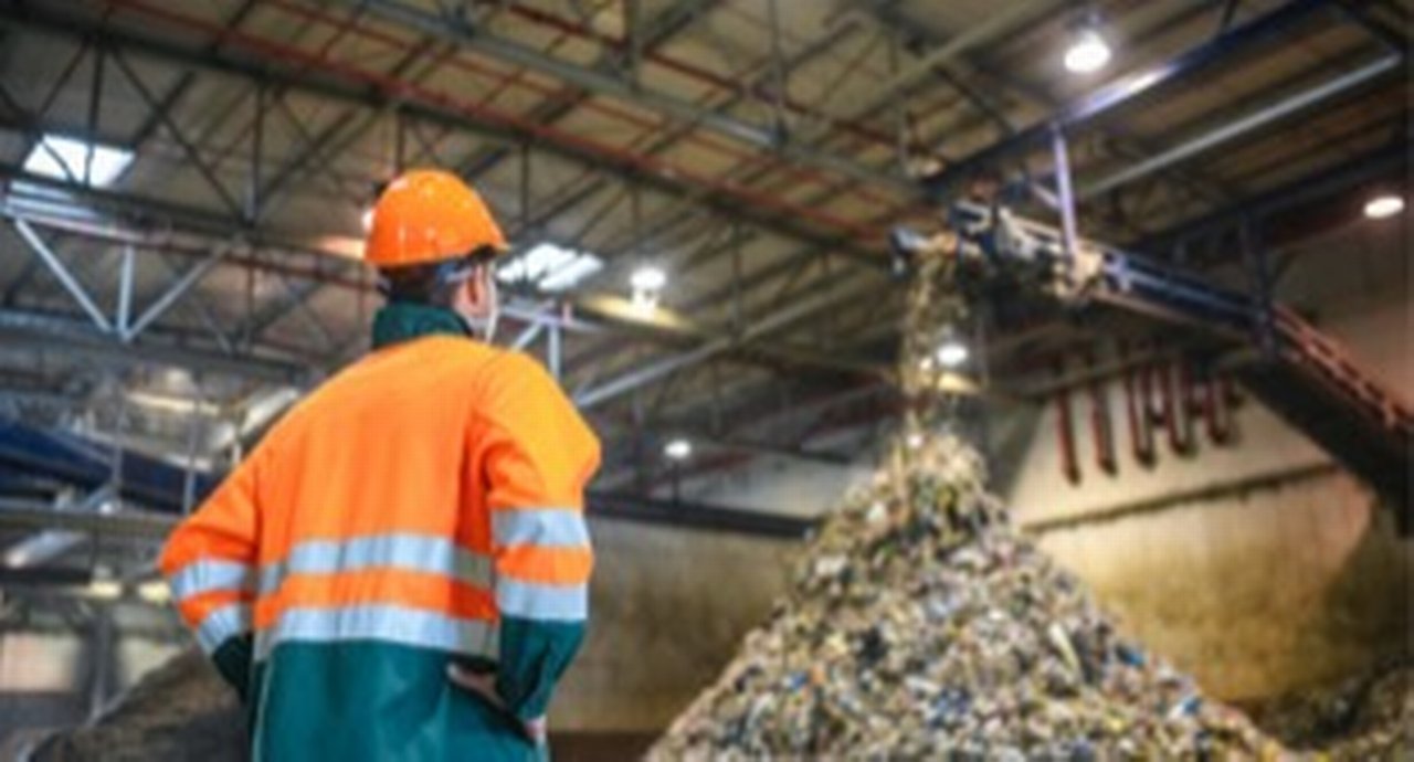 Low angle rear view of young male worker in helmet, pollution mask, and reflective clothing observing waste falling from conveyor belt onto pile at facility