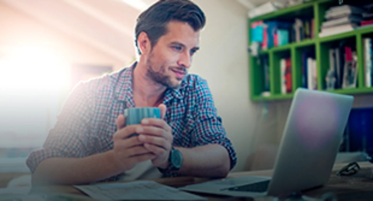 A young man in front of his laptop holding a cup in his hands