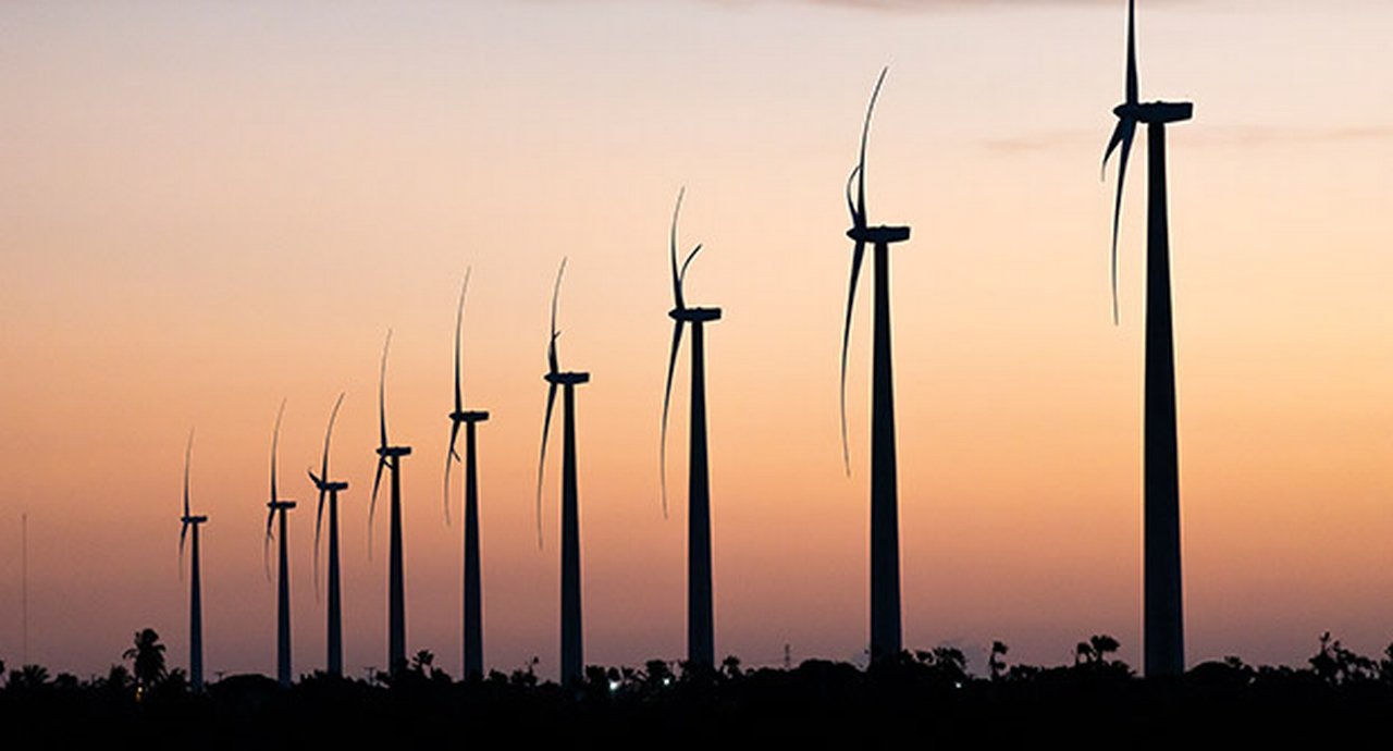 Nine wind turbines in a row against an orange sky at dusk in São Miguel do Gostoso, Rio Grande do Norte, Brazil.