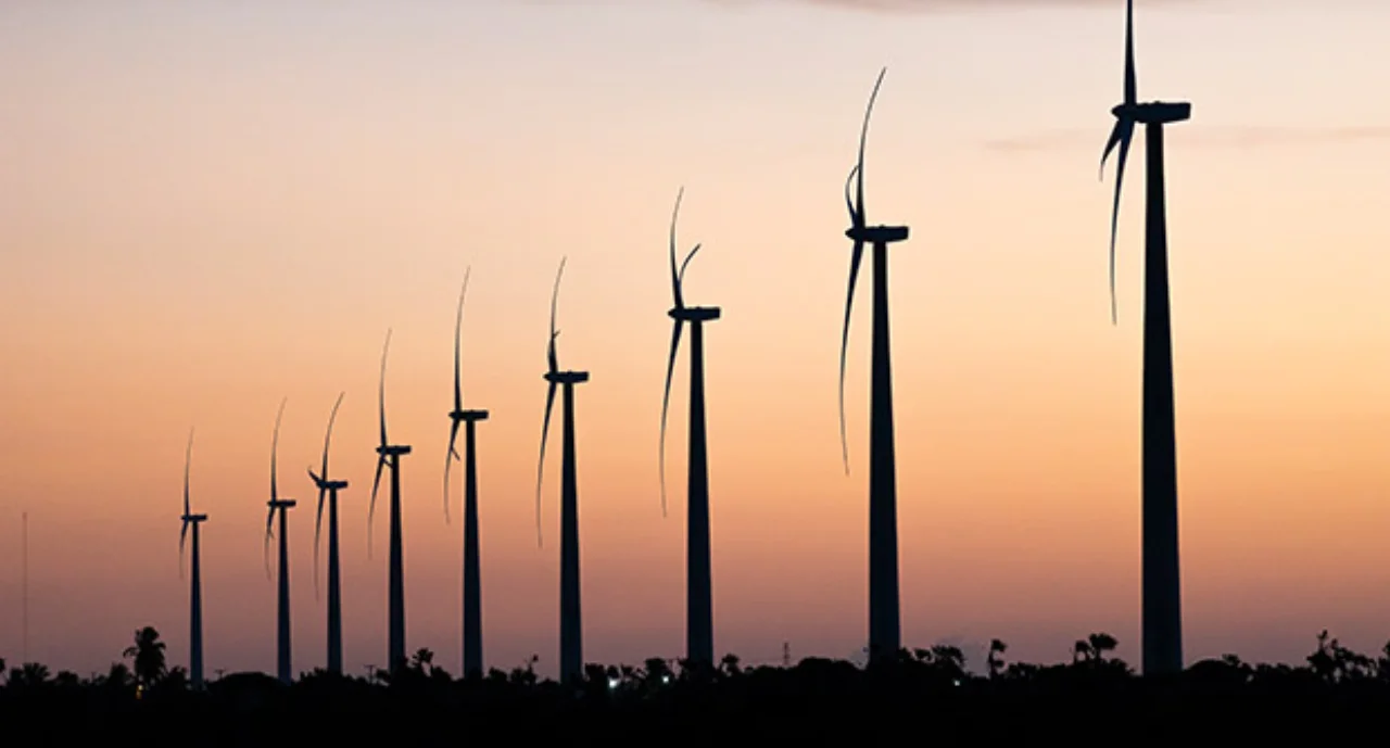 Nine wind turbines in a row against an orange sky at dusk in São Miguel do Gostoso, Rio Grande do Norte, Brazil.