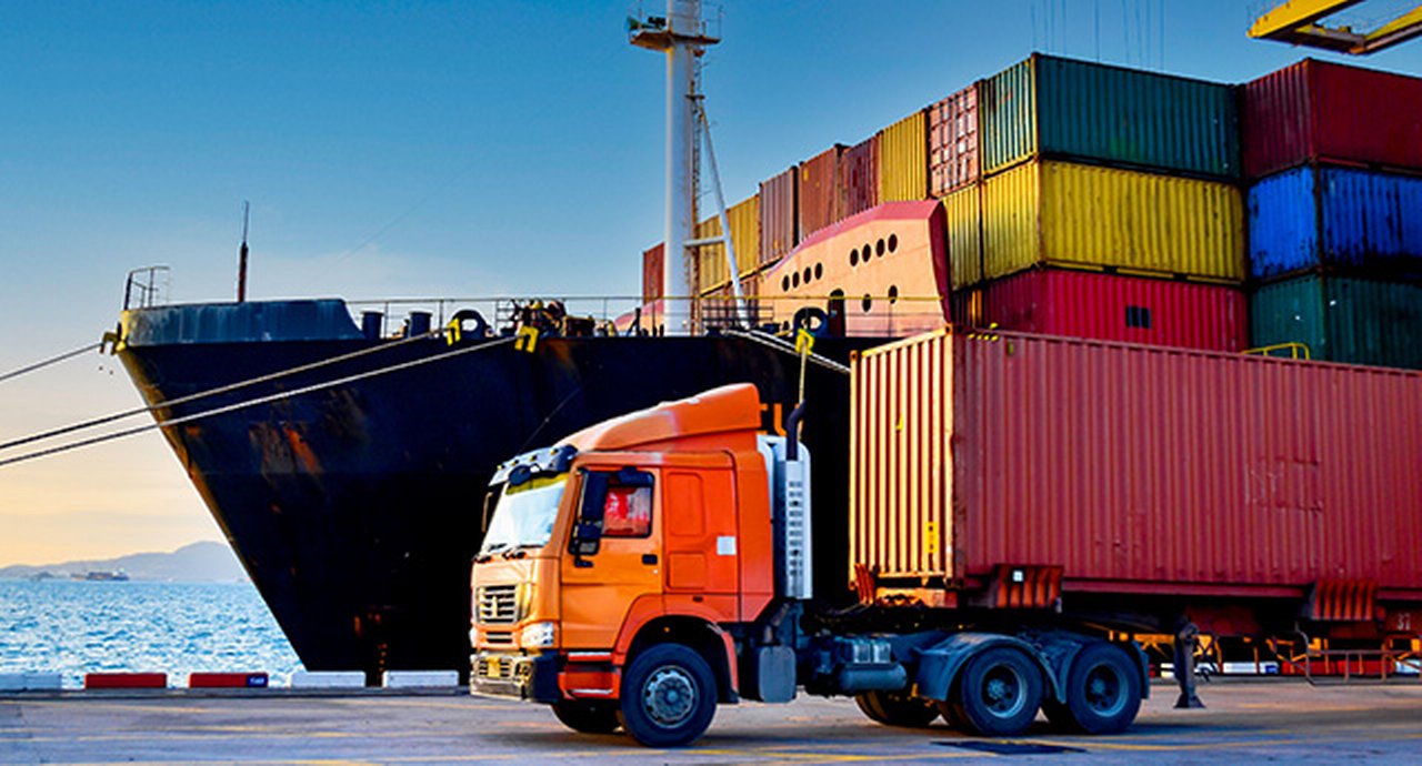 Truck carrying forty-foot container leaving port terminal with ship and quay crane on the background