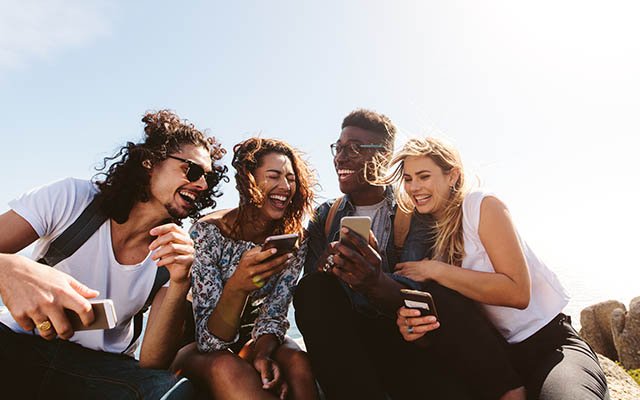 Young group of people sitting on the top of the mountain, using smartphones and smiling. Different friends enjoying a day out.