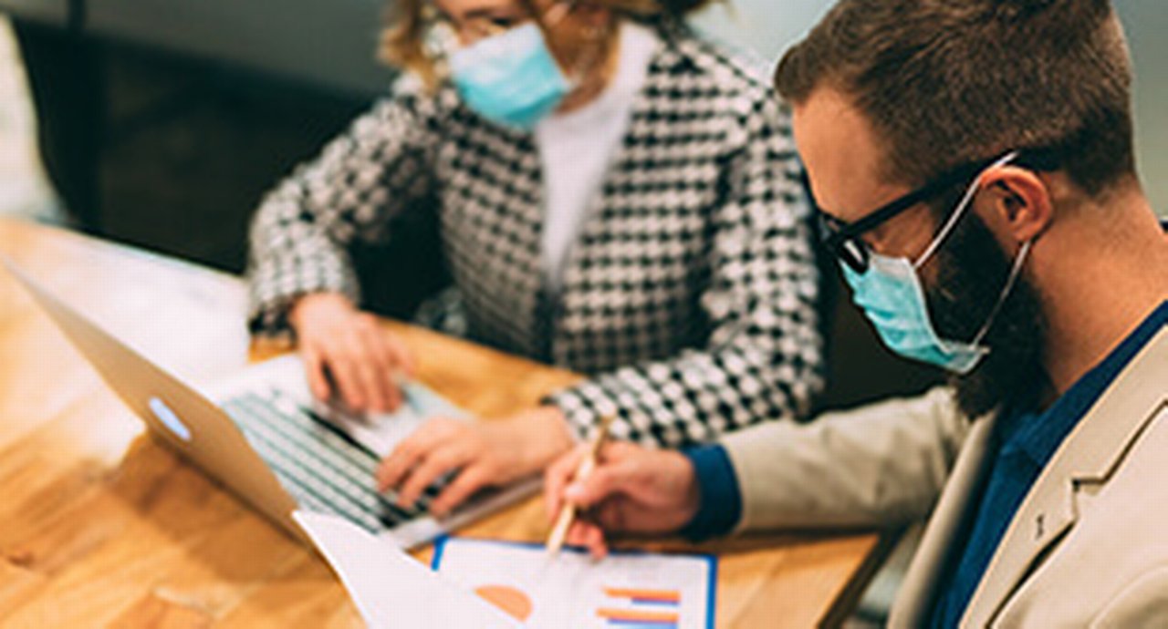 a woman and a man wearing masks sitting in front of a laptop
