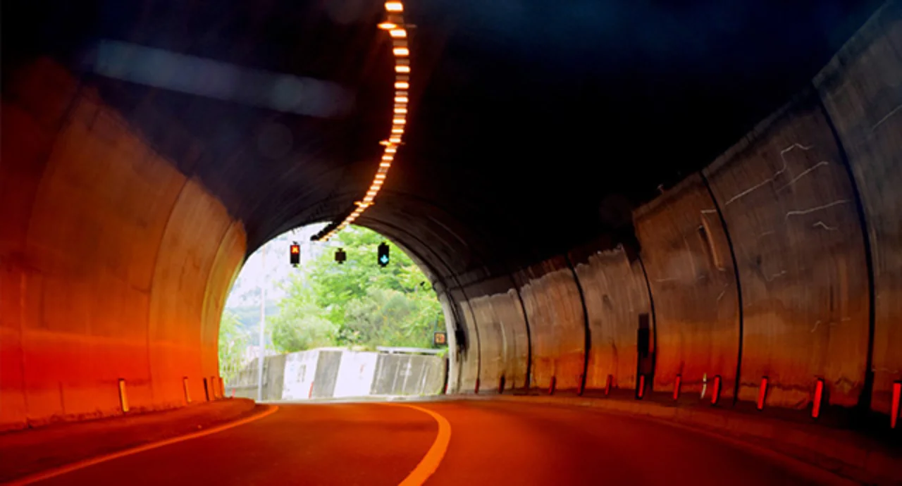 A curved road tunnel lit by warm orange ceiling lights, with a bright opening at the far end revealing green trees and daylight, and traffic signals hanging above the exit.
