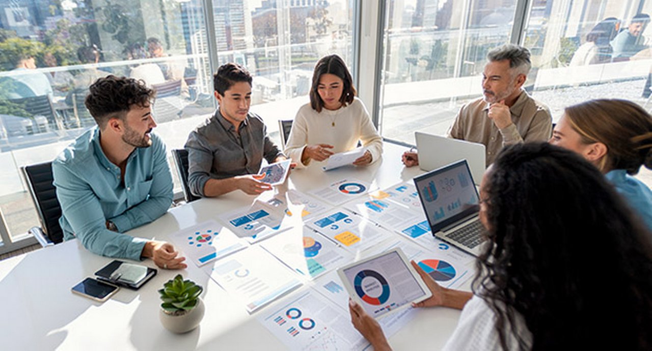 Group of people working with paperwork at a boardroom table during a business presentation or seminar. The documents contain financial or marketing figures, graphs and charts.