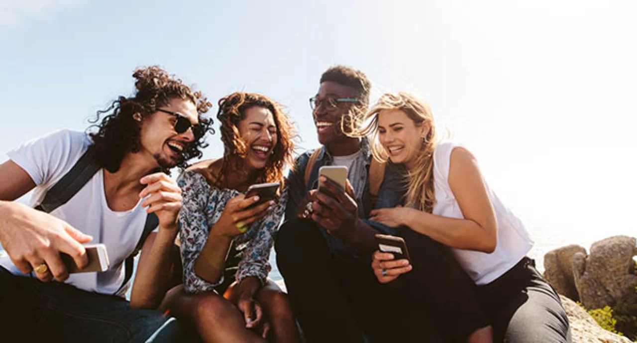 Young group of people sitting on the top of the mountain, using smartphones and smiling. Different friends enjoying a day out.