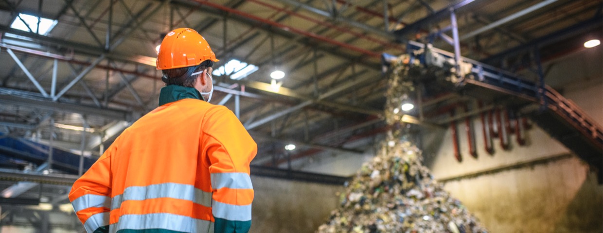 Low angle rear view of young male worker in helmet, pollution mask, and reflective clothing observing waste falling from conveyor belt onto pile at facility