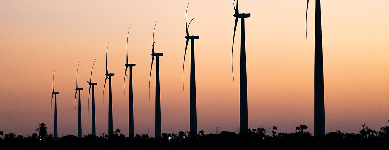 Nine wind turbines in a row against an orange sky at dusk in São Miguel do Gostoso, Rio Grande do Norte, Brazil.