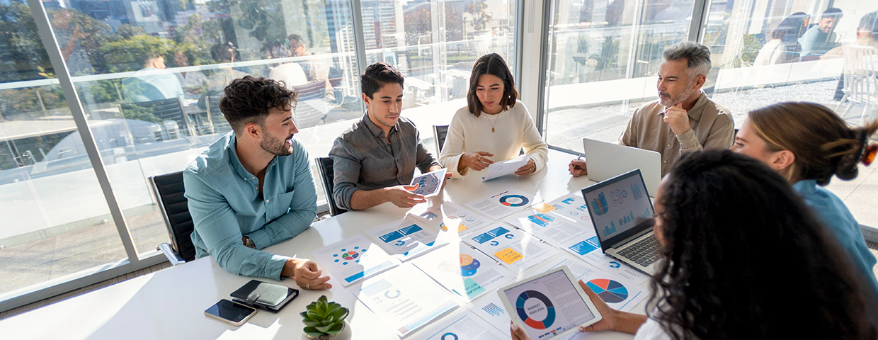 Group of people working with paperwork at a boardroom table during a business presentation or seminar. The documents contain financial or marketing figures, graphs and charts.