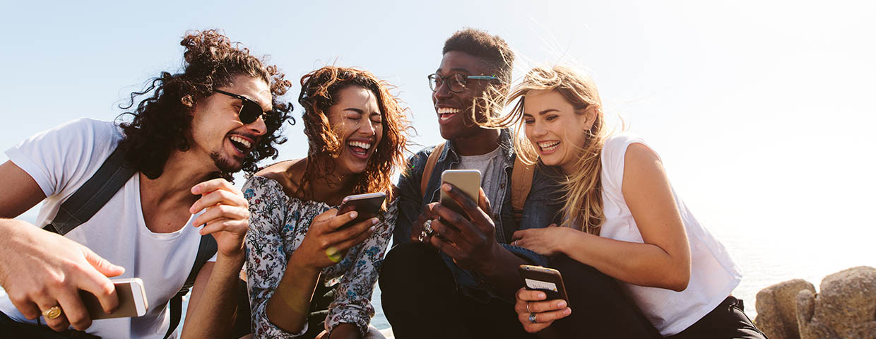 Young group of people sitting on the top of the mountain, using smartphones and smiling. Different friends enjoying a day out.